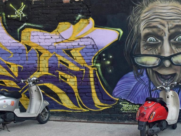 Ein Graffiti mit dem Titel "Verrückte Oma" zeigt an einer Hauswand eine alte Frau, davor stehen mehrere Motorroller.