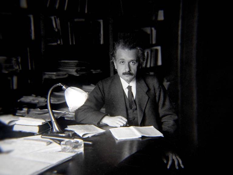 Albert Einstein sitzt an einem Schreibtisch, auf dem eine Lampe brennt, im Hintergrund Bücherregale
