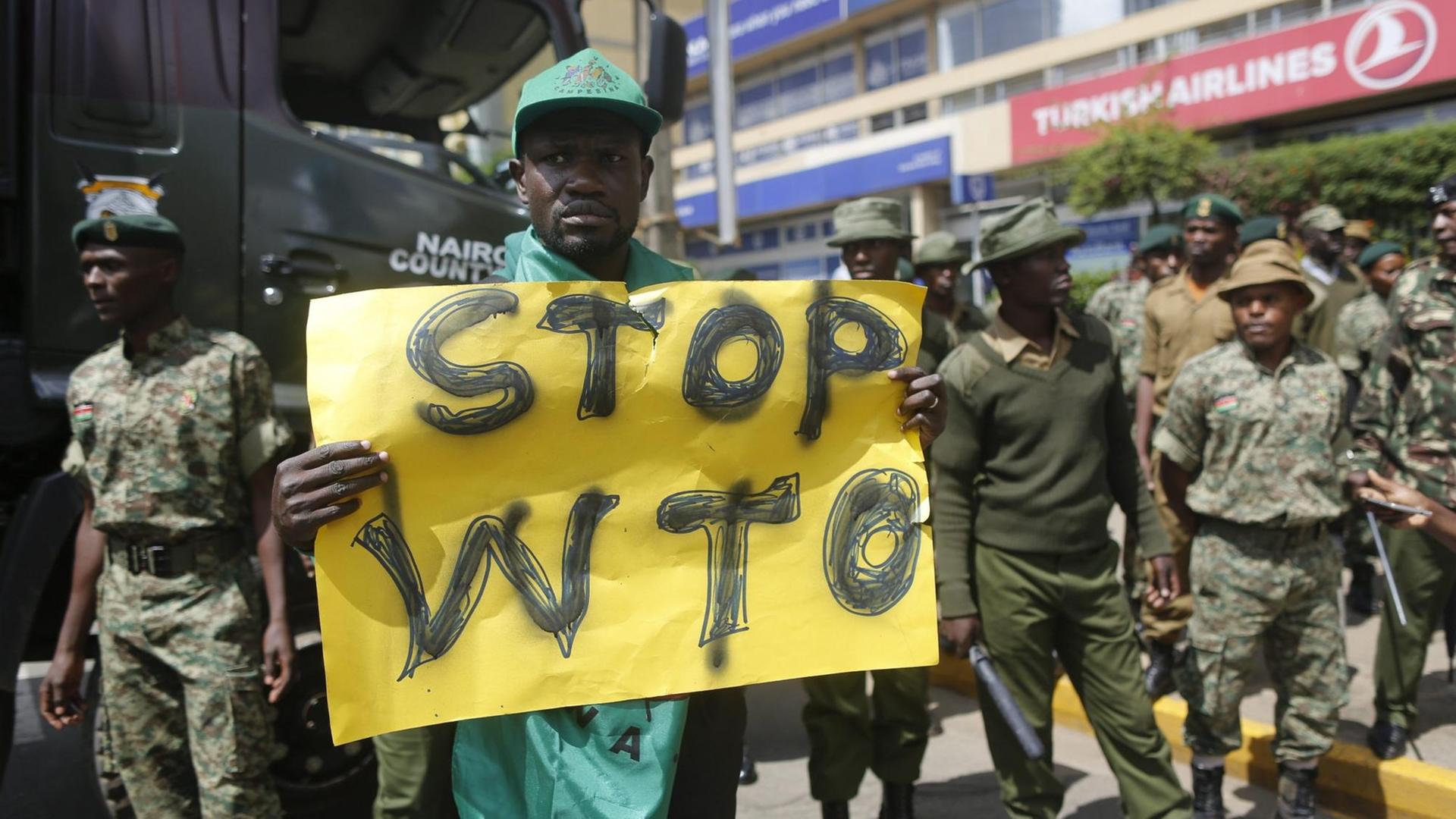Ein kenianischer Aktivist hält ein Transparent hoch, auf dem "Stop WTO" zu lesen ist. Um ihn herum stehen Sicherheitsleute.