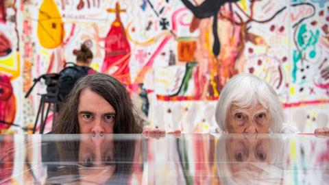 Brigitte und Jonathan Meese (Maler und Aktionskünstler) spiegeln sich im Ausstellungsraum in einem Schaukasten. Im Hintergrund sind Gemälde von Jonathan Meese zu sehen.