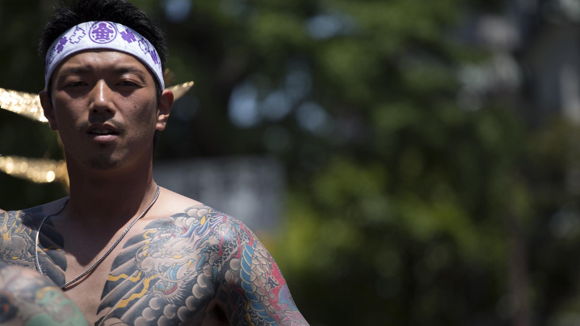 Ein junger Japaner zeigt seinen großflächig tätowierten Oberkörper. Er hat sich ein weißes Band mit lila Zeichen um den Kopf gebunden.