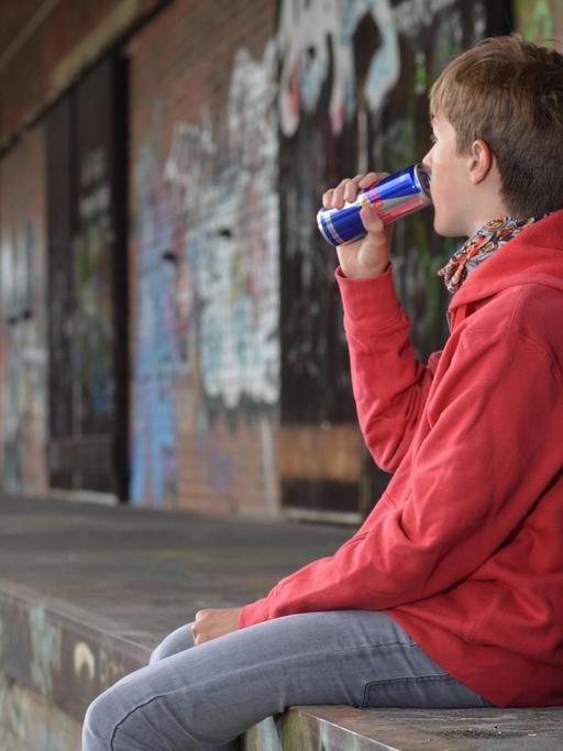 Ein Jugendlicher sitzt auf einer Steinmauer und trinkt einen Energy Drink.