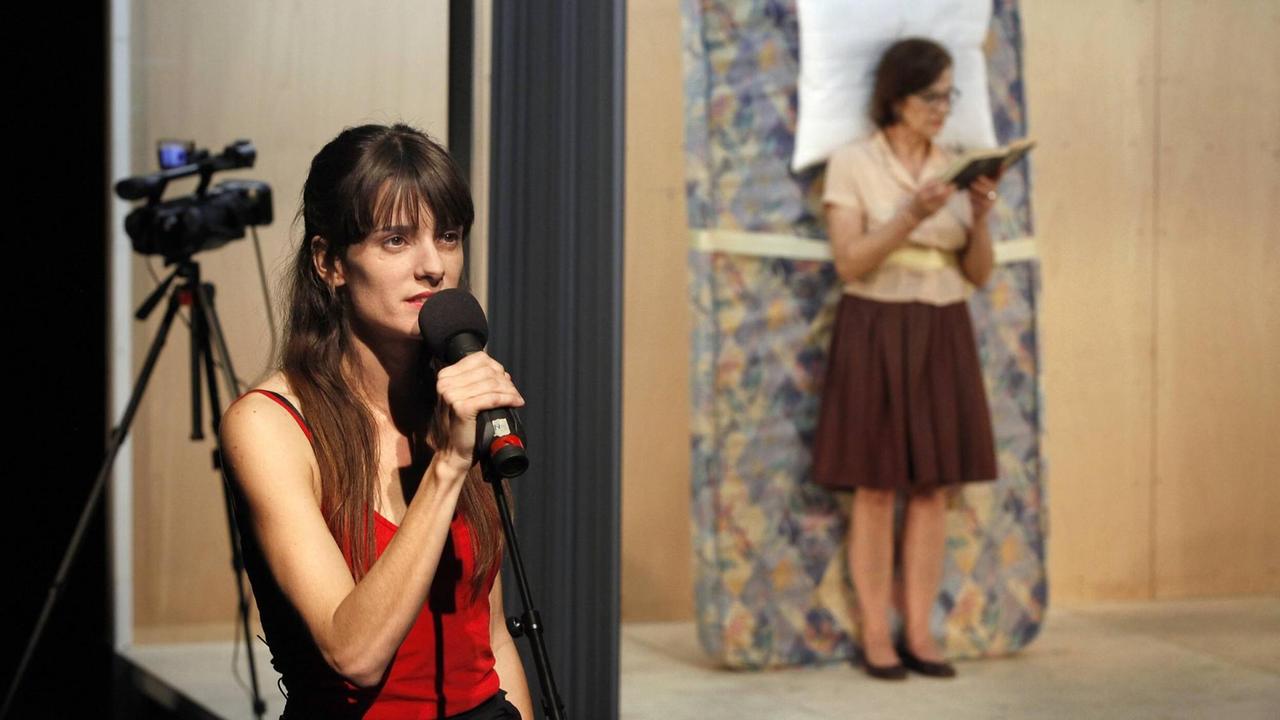 Lola Arias spricht auf Bühne in ein Mikrofon. Im Hintergrund eine Kamera und ihre Schauspielkollegin Elvira Onetto, die vor einer Matratze steht und ein Buch liest.