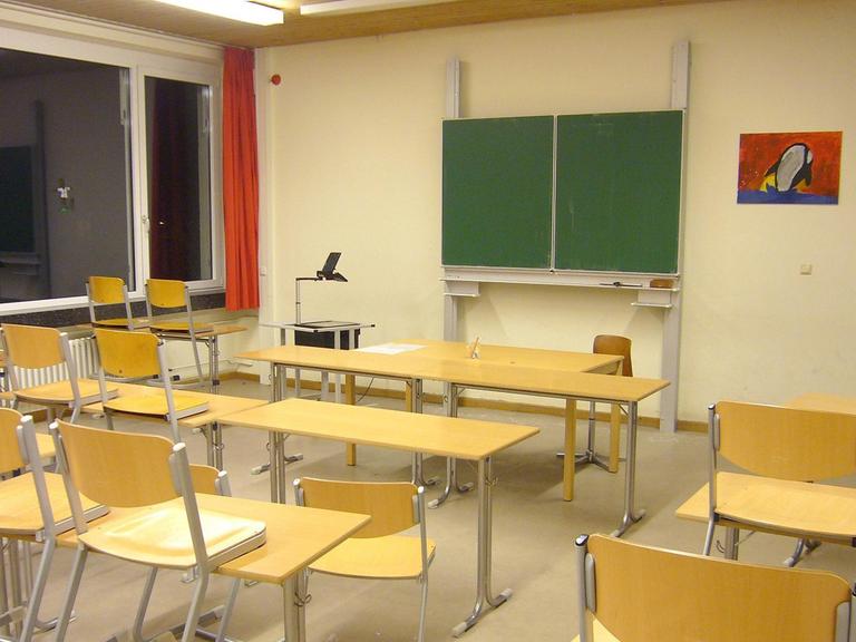 Ein leeres Klassenzimmer. Die Tafel ist abgewischt und zugeklappt. Die Stühle aus hellbraunem Holzfurnier stehen auf den Tischen.