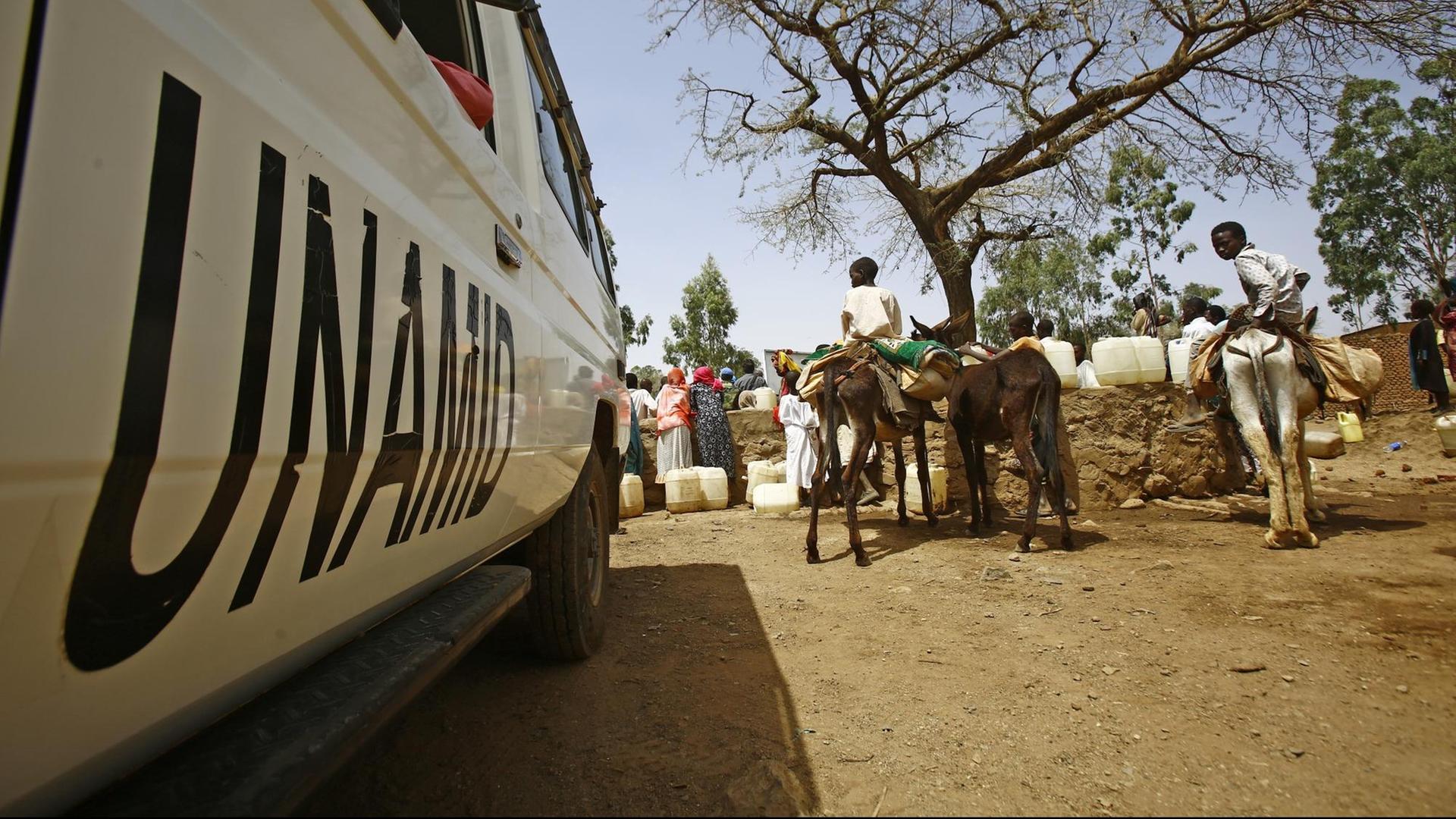Ein Fahrzeug mit dem Aufdruck UNAMID steht auf einem sandigen Platz. Daneben stehen Frauen mit Wasserkanistern. Kinder sitzen auf bepackten Eseln.