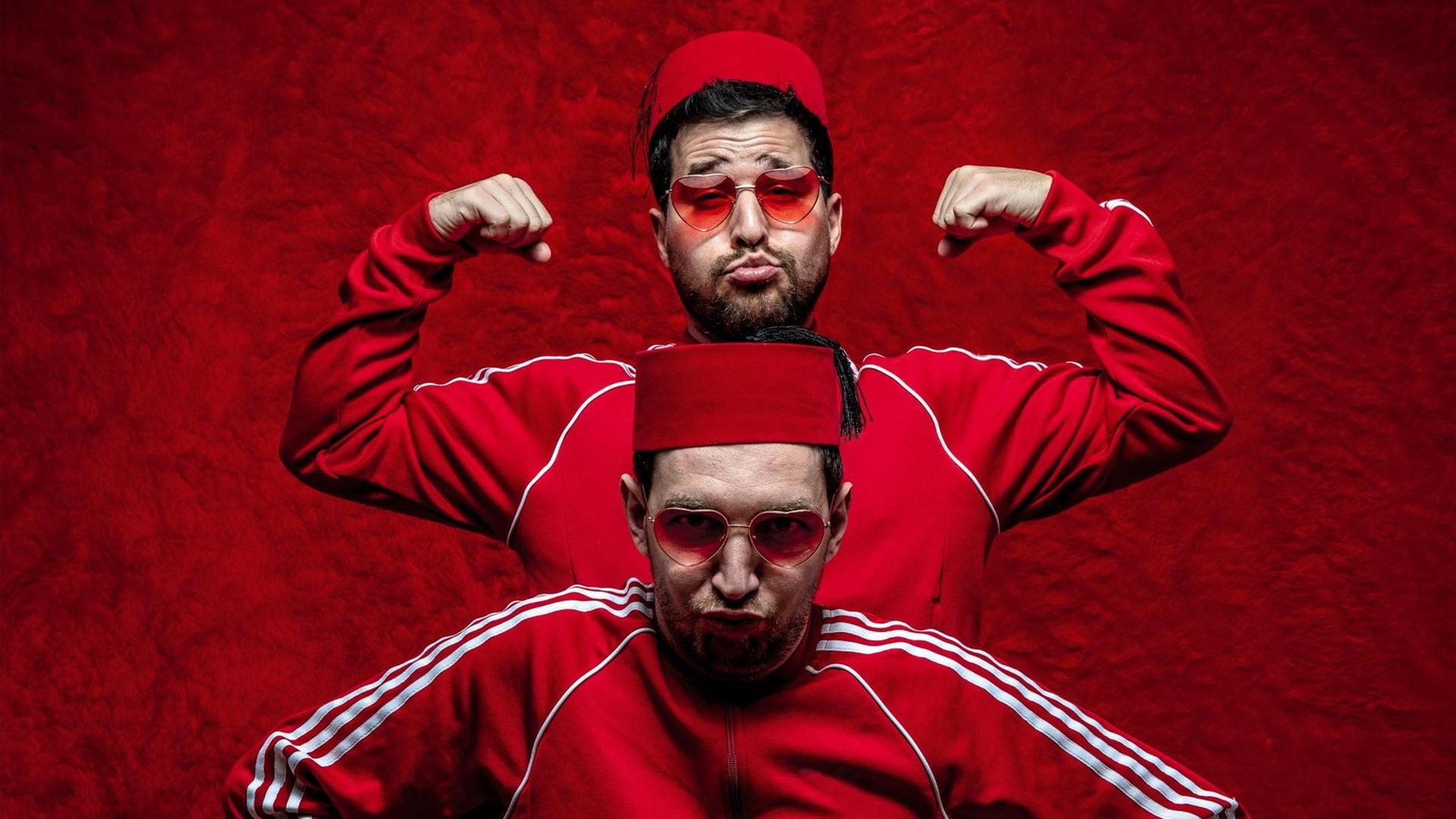 Zu sehen sind zwei Männer, die hintereinanderstehen. Sie tragen einen Fes auf dem Kopf und rote Adidas-Trainingsanzüge.