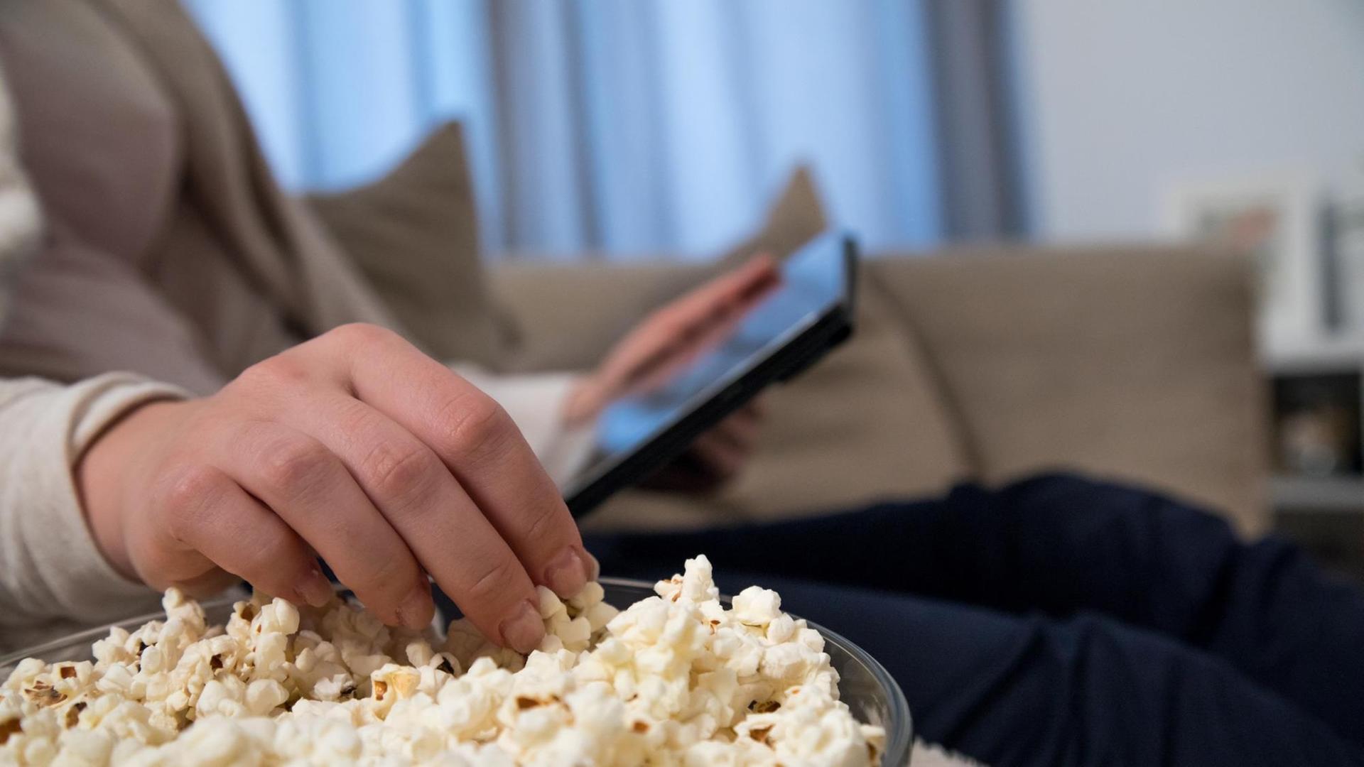 Eine junge Frau sitzt mit Popcorn auf der Couch und schaut auf ihrem Tablet einen Film.