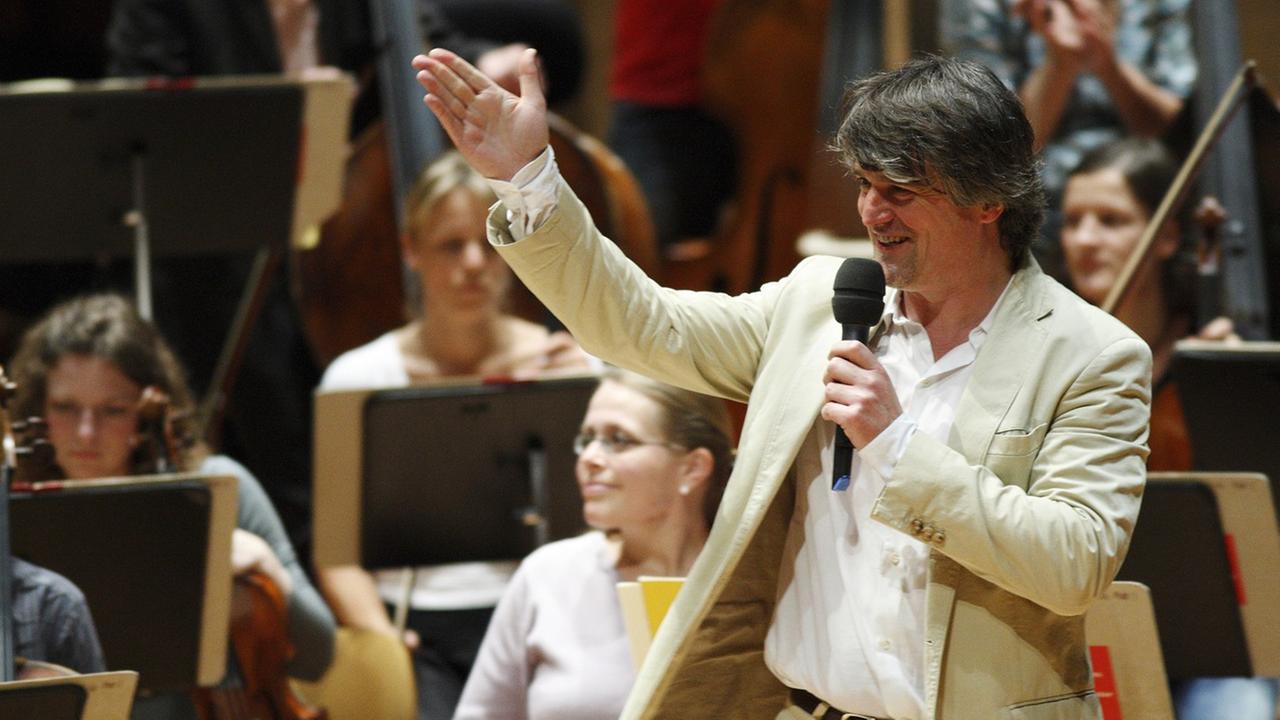 Der Dirigent Ingo Metzmacher im hellen Anzug zeigt lachend Richtung Musiker des DSO Berlin.