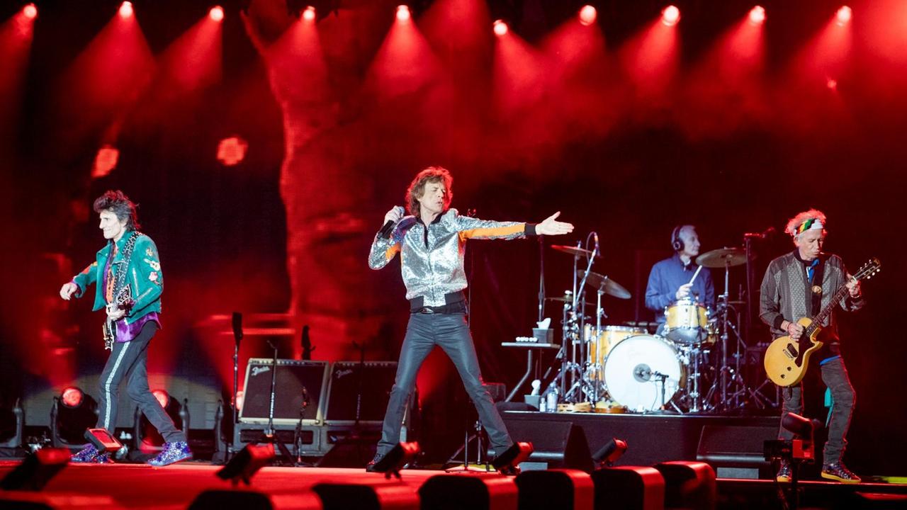 Drei Männer mit Musikinstrumenten und ein Mann mit Mikrofon in der Hand stehen auf einer rot ausgeleuchteten Bühne.