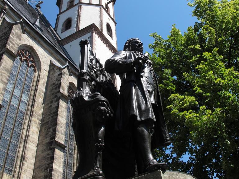 Aus der Froschperspektive fotografiert: das Denkmal von Johann Sebastian Bach. Im Hintergrund die Thomaskirche und ein großer Baum mit grünen Blättern.