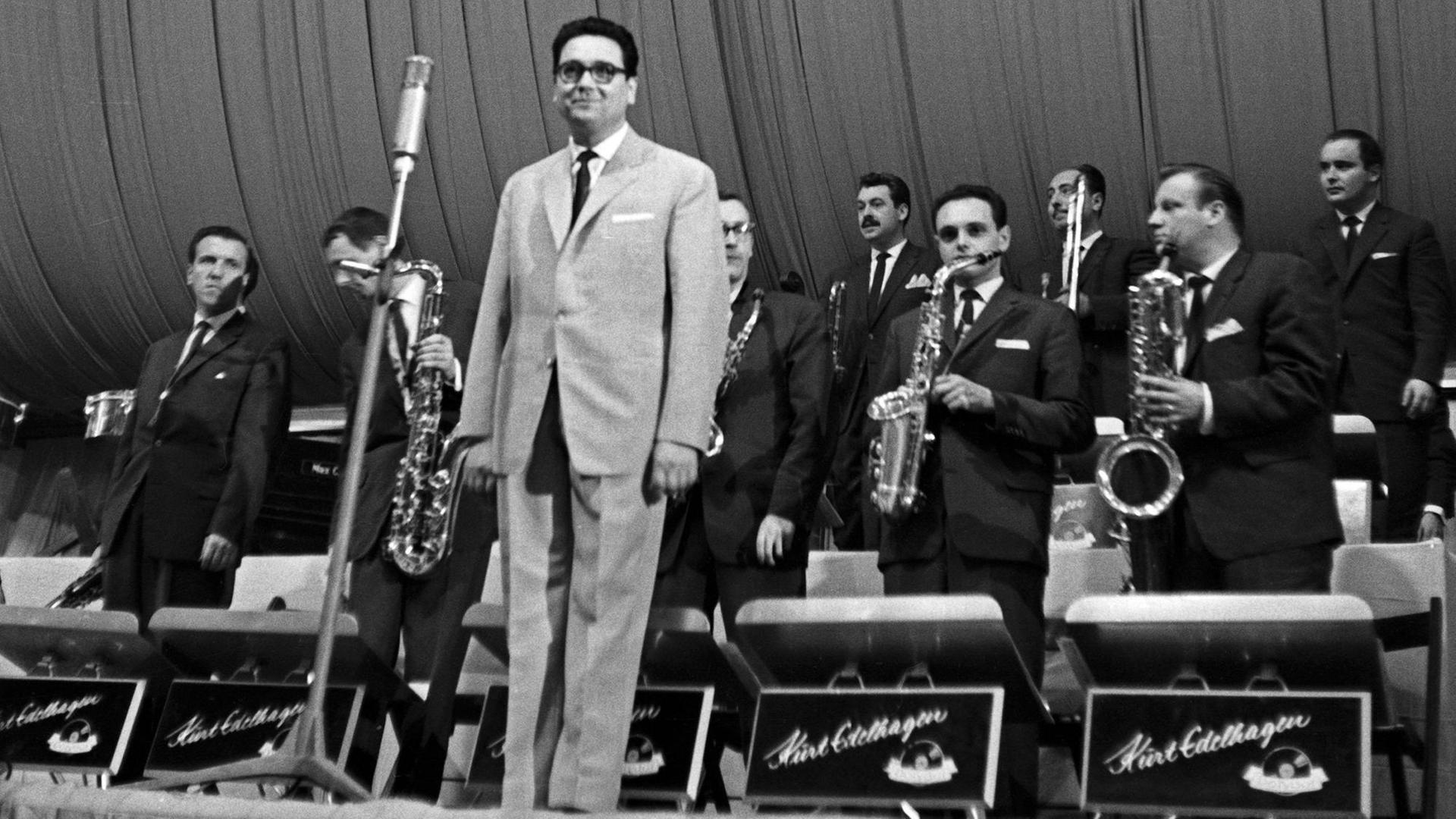 Ein altes s/w Bild, vermutlich 1950er oder 1960er Jahre. Ein Orchesterleiter im Anzug steht zum Publikum gewendet lächelnd vor der ebenfalls stehenden Band. Auf den Pulten der Bläser der Schriftzug "Kurt Edelhagen".