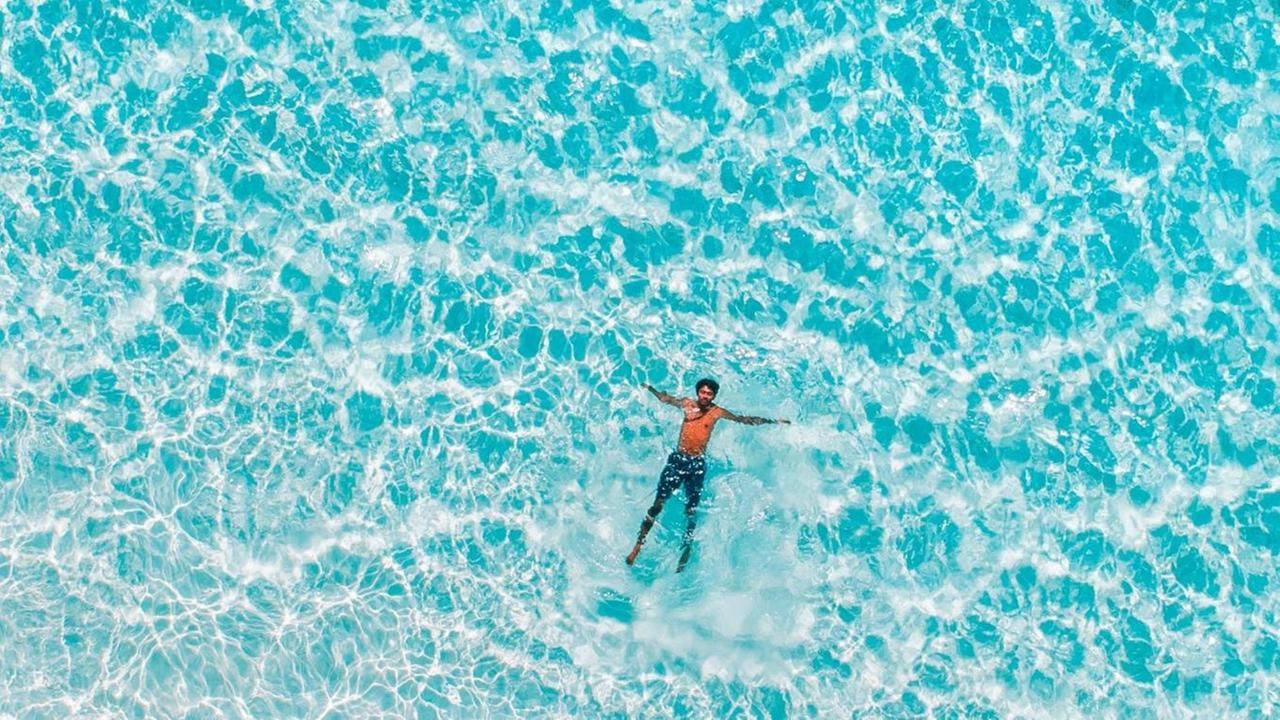 Vogelperspektive auf einen Mann, der im türkisblauen Meer schwimmt.