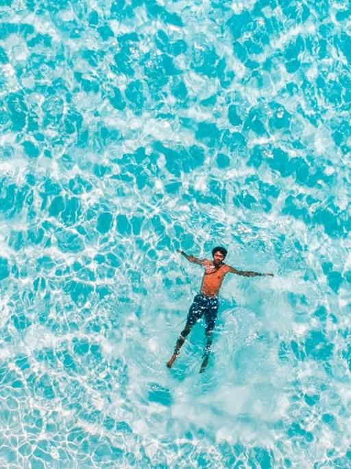 Vogelperspektive auf einen Mann, der im türkisblauen Meer schwimmt.