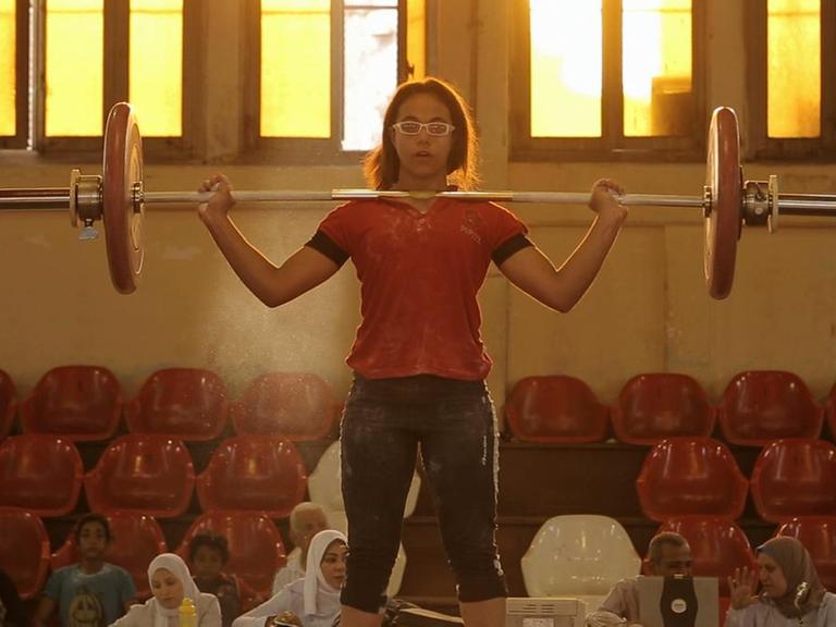 Eine Frau beim Gewichtheben - angefeuert von vielen Frauen im Hintergrund.