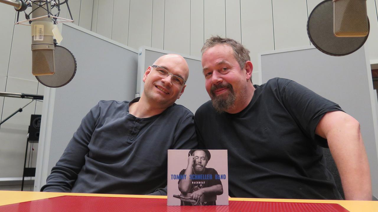 Zwei Männer sitzen nebeneinander an einem Tisch und lächeln in die Kamera.