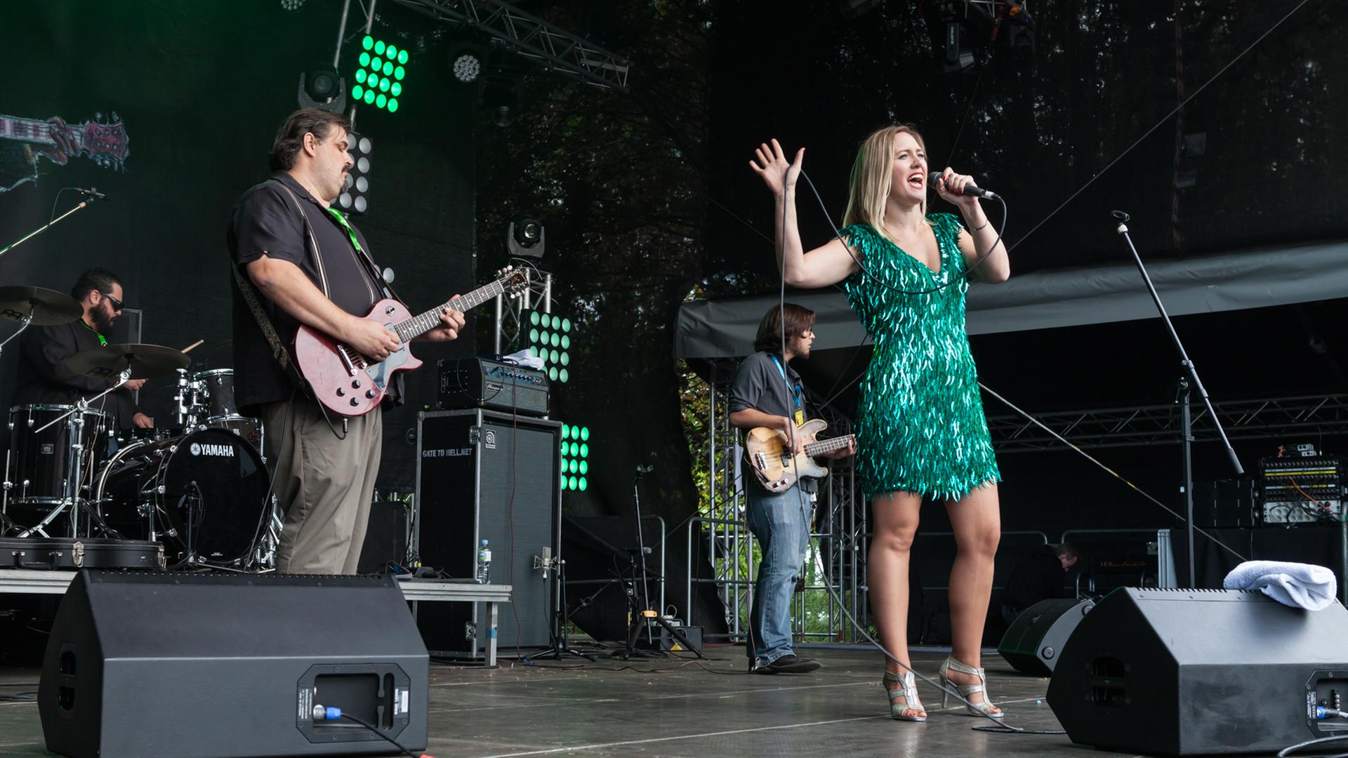 Eine blonde Frau steht in einem grünen Paillettenkleid auf der Bühne und singt, im Hintergrund spielt ihre Band.