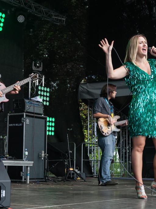 Eine blonde Frau steht in einem grünen Paillettenkleid auf der Bühne und singt, im Hintergrund spielt ihre Band.