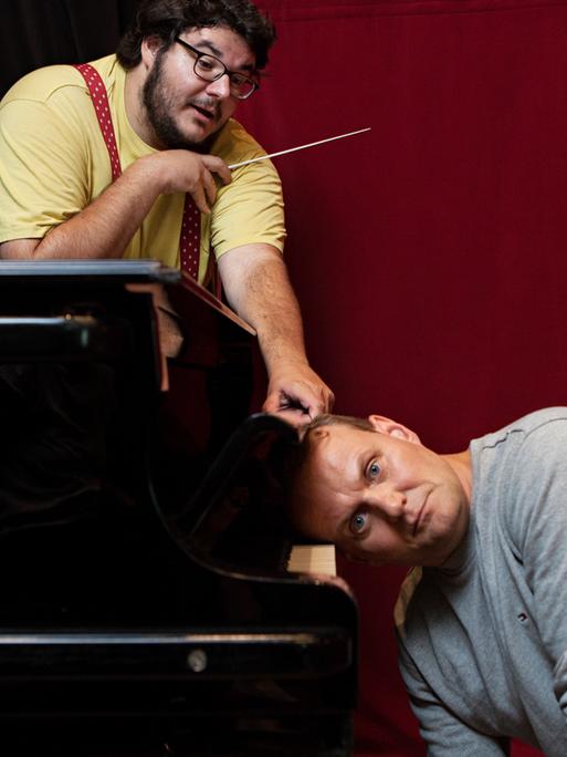 Das Bild zeigt Axel Ranisch, wie er hinter einem Klavier mit einem Dirigentenstab nach rechts zeigt, während Devid Striesow mit dem Kopf auf den Tasten des Klaviers liegt.