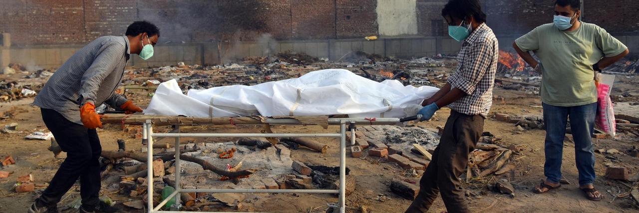 In Neu-Delhi schieben zwei Männer eine Bahre mit einem Toten an einen Platz, auf dem Leichen verbrannt  werden.