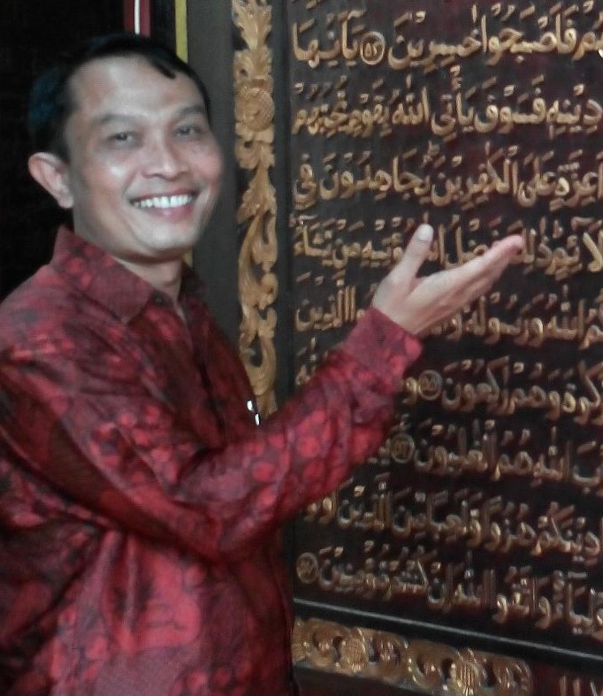 Sahiron Syamsuddin zeigt auf eine schwarze Tafel, auf der in goldener arabischer Schrift ein Koranauszug abgebildet ist.