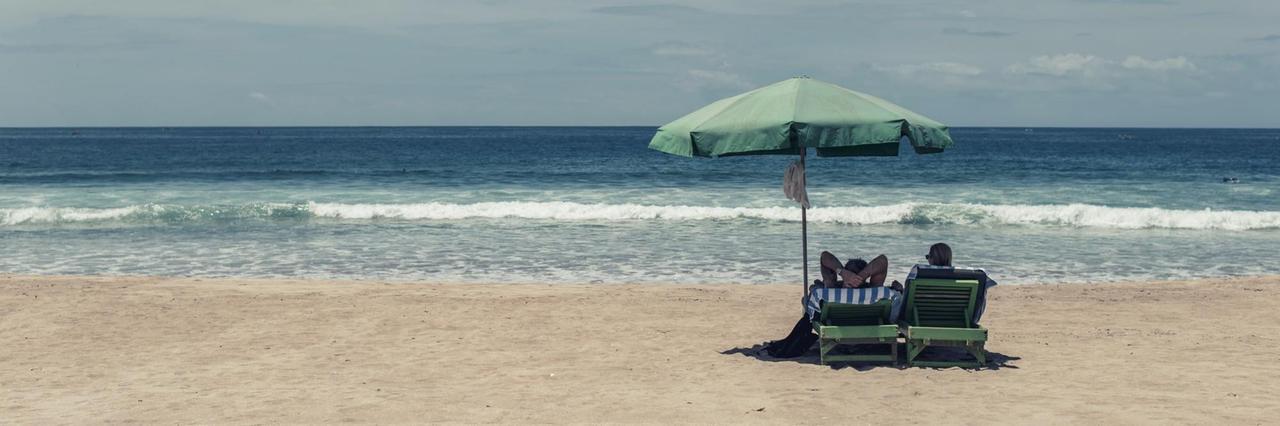 Bild eines fast komplett leeren Strands mit Blick aufs Meer: Nur zwei Menschen liegen auf ihren Strandliegen unter einem Sonnenschirm. Bild eines fast komplett leeren Strands mit Blick aufs Meer: Nur zwei Menschen liegen auf ihren Strandliegen unter einem Sonnenschirm.