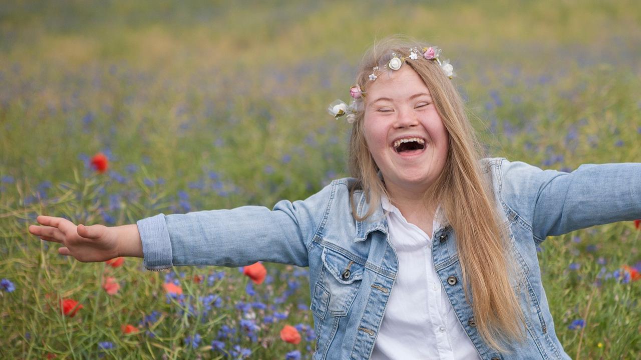 Eine fröhliche junge Frau steht in einem Garten und blickt mit ausgebreiteten Armen in die Kamera. Auf dem blonden Haar trägt sie einen Blumenkranz.