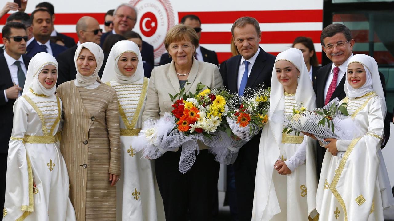 Sie sehen Bundeskanzlerin Merkel und EU-Ratspräsident Tusk in einem Flüchtlingslager in der Türkei. Sie sind umringt von Frauen in hellen Trachten.