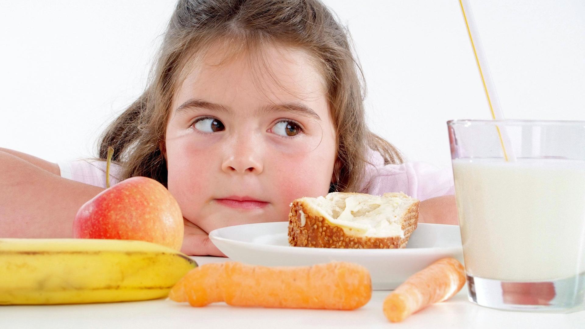 Ein Mädchen sitzt vor Obst, Gemüse, Brot und Milch - Lebensmittel die gemeinhin als gesund gelten