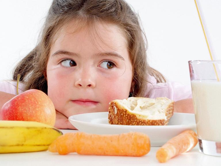 Ein Mädchen sitzt vor Obst, Gemüse, Brot und Milch - Lebensmittel die gemeinhin als gesund gelten