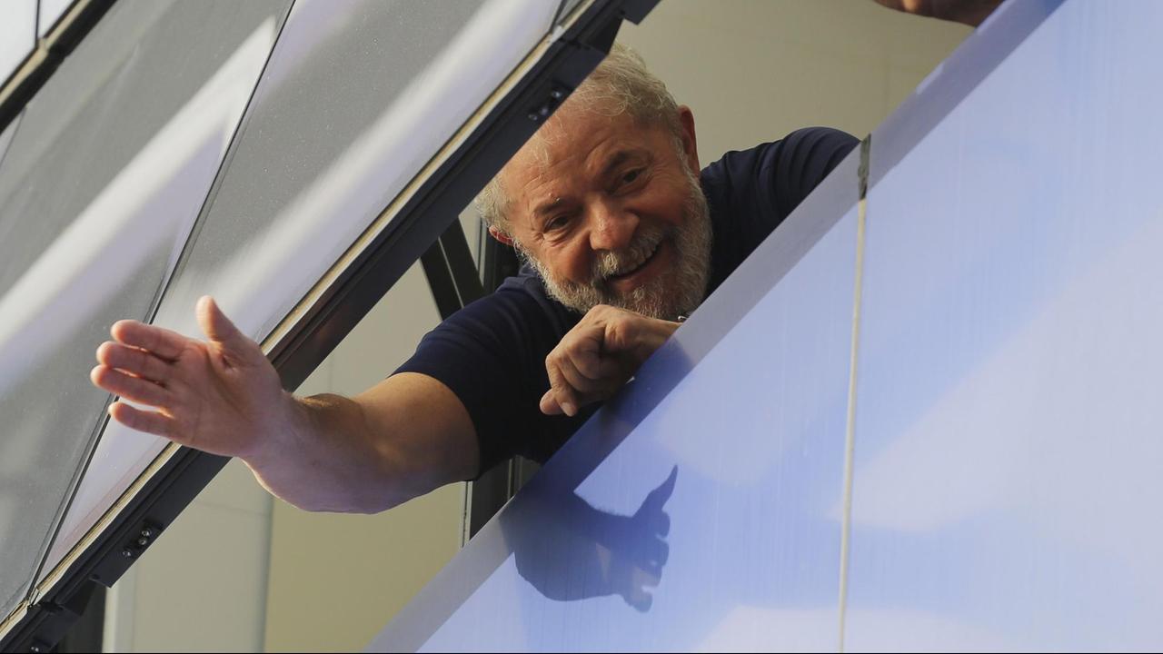 Lula winkt aus einem gekippten Fenster lächelnd nach unten. Daneben sieht man die Hand einer weiteren Person.