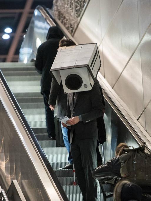 Ein Mann im Anzung und mit einer Überwachungskamera aus Pappe auf dem Kopf fährt eine Rollpreppe hoch.