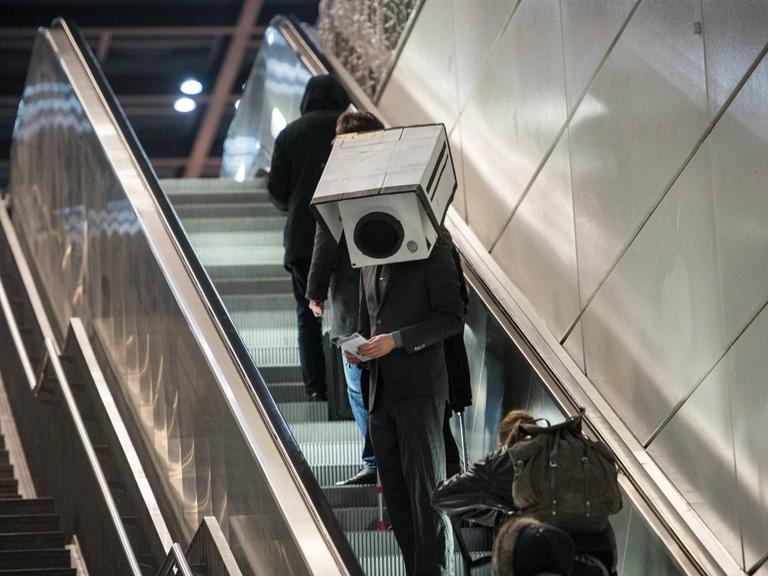 Ein Mann im Anzung und mit einer Überwachungskamera aus Pappe auf dem Kopf fährt eine Rollpreppe hoch.
