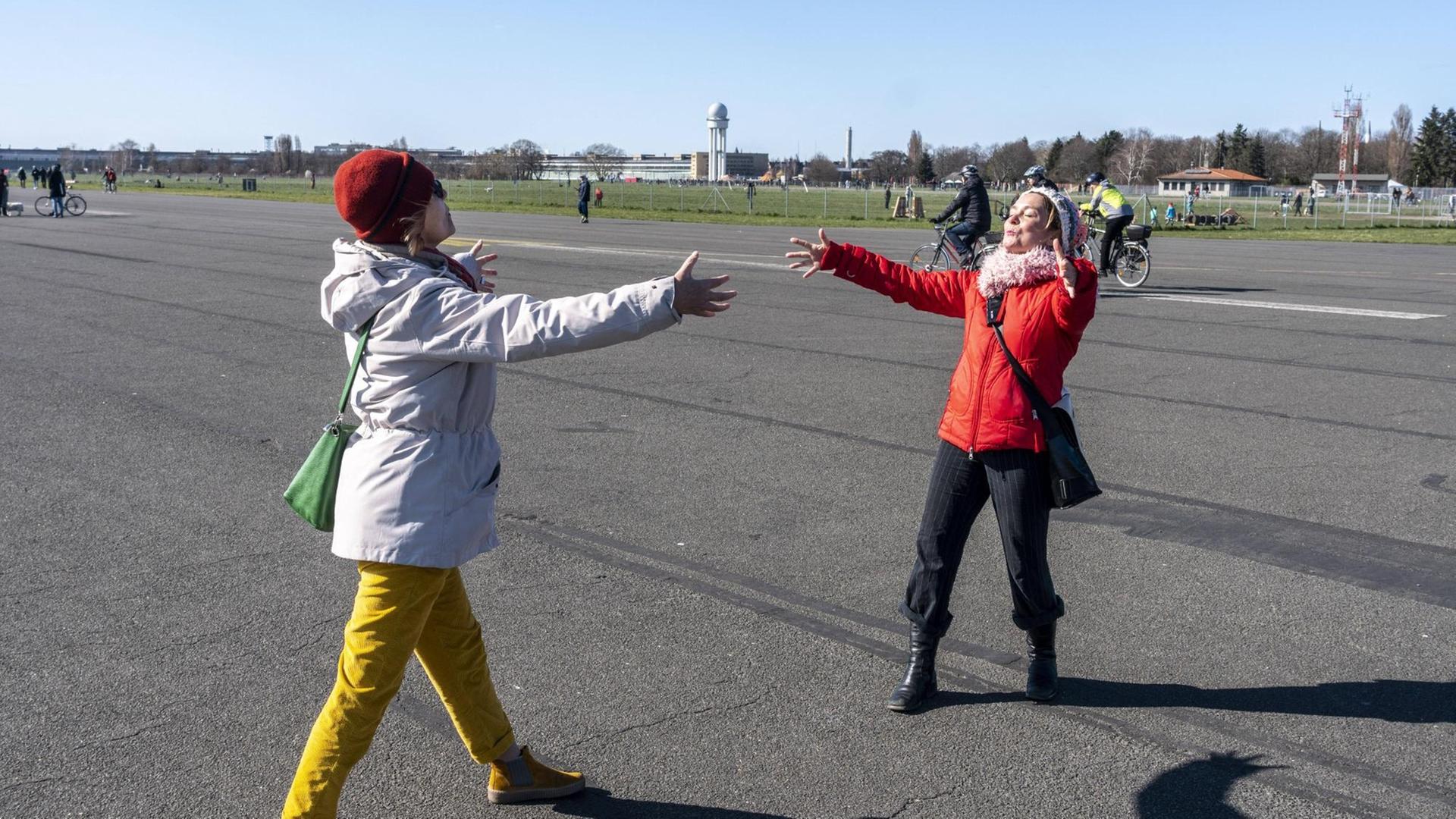 Zwei Frauen Begrüßen sich mit einer angedeuteten Umarmung aus der Distanz auf dem Tempelhofes Feld in Berlin.
