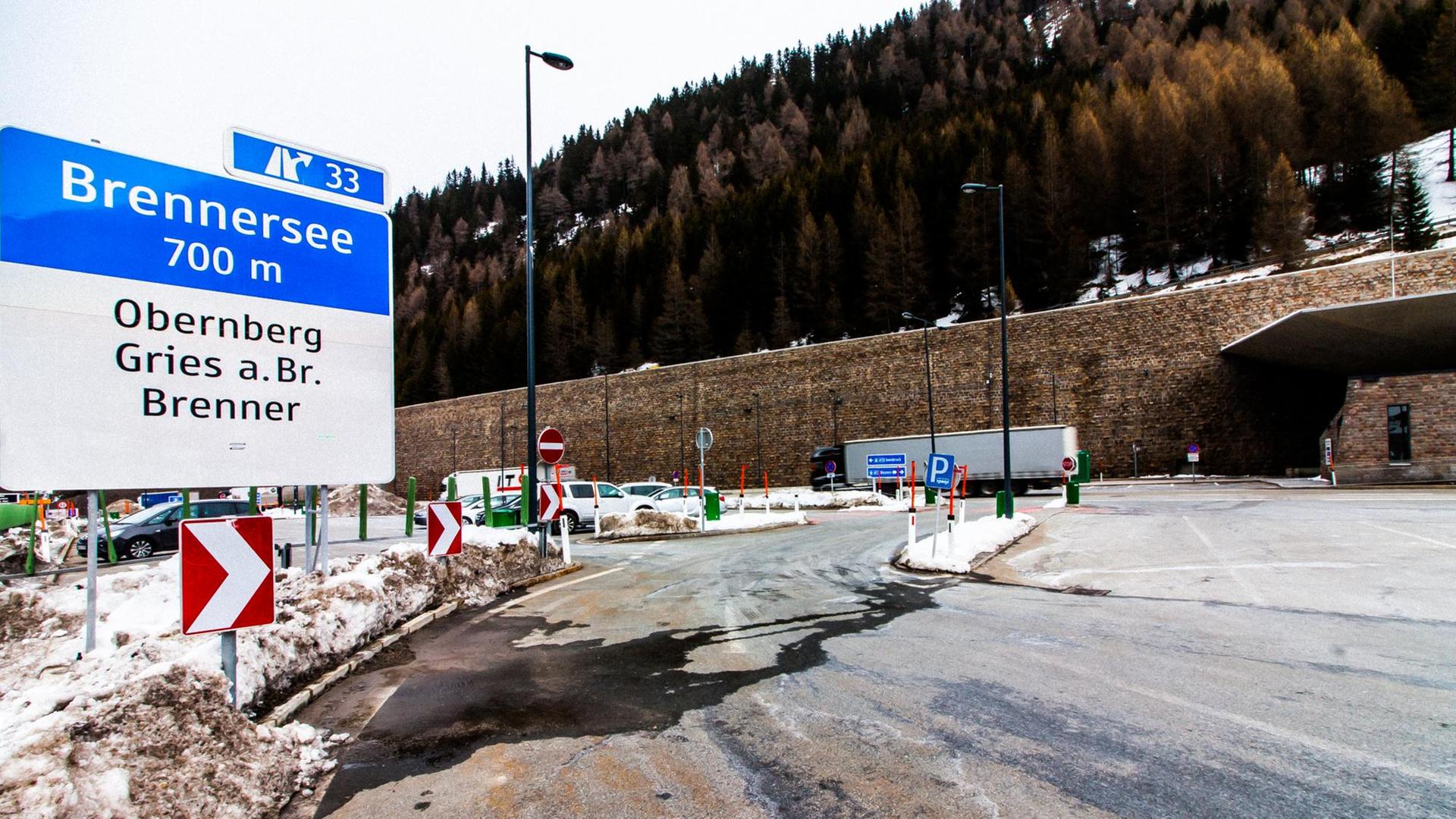 Lkws befahren den Brennerpass. Im Hintergrund die schneebedeckte Tannenwälder, im Vordergrund sieht man ein Schilder Richtung Brennersee und Obernberg Gries am Brenner