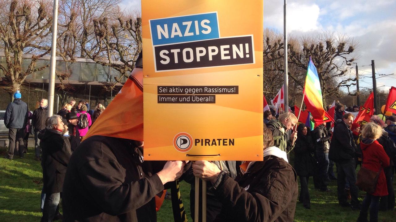 Demonstranten halten ein Schild der Piraten-Partei mit der Aufschrift "Nazis stoppen - Sei aktiv gegen Rassismus! Immer und Überall!" Im Hintergrund sind zahlreiche weitere Demonstranten unter anderem mit Gewerkschafts- und Regenbogenflaggen zu sehen.