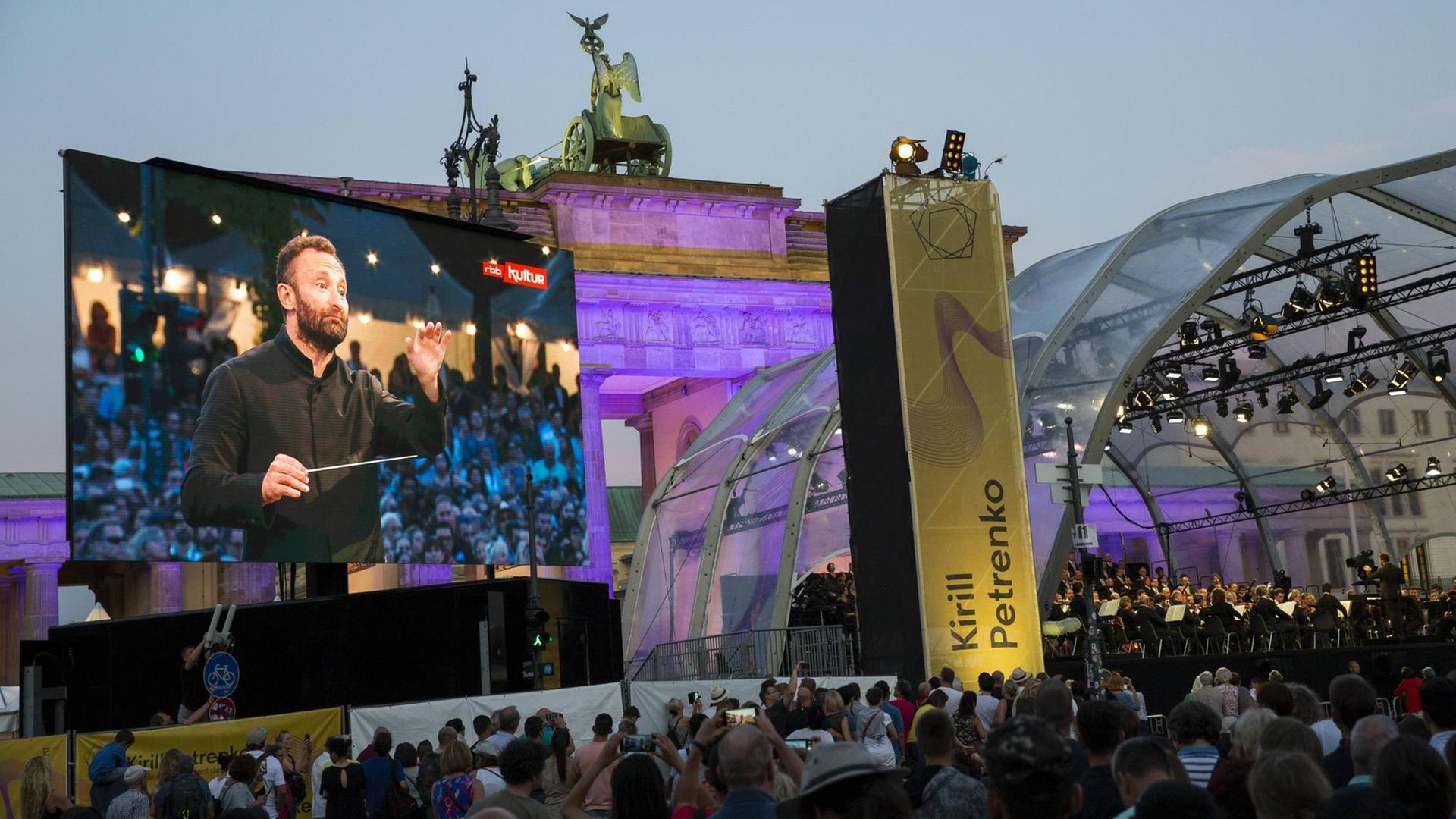 Kirill Petrenko ist auf einer großen Leinwand dabei zu sehen, wie er die Berliner Philharmoniker dirigiert, im Hintergrund das Brandenburger Tor.