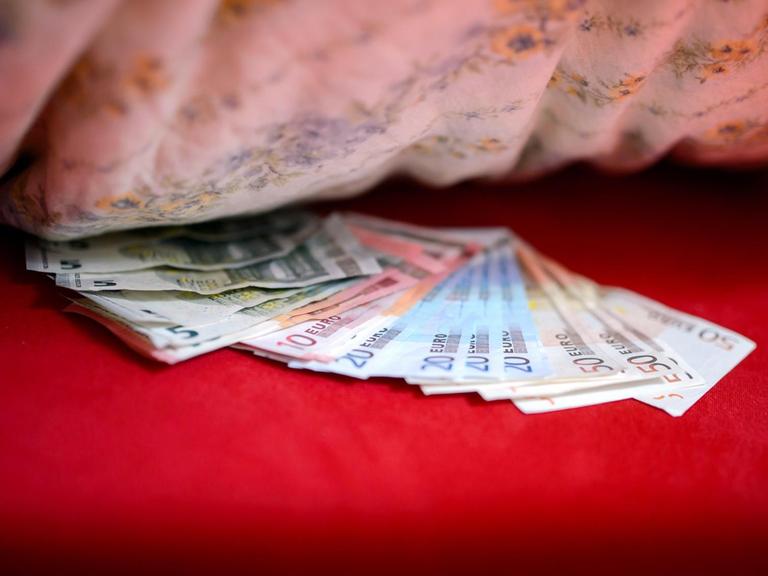Man sieht Geldscheine auf einer roten Bettdecke unter einem Kopfkissen.