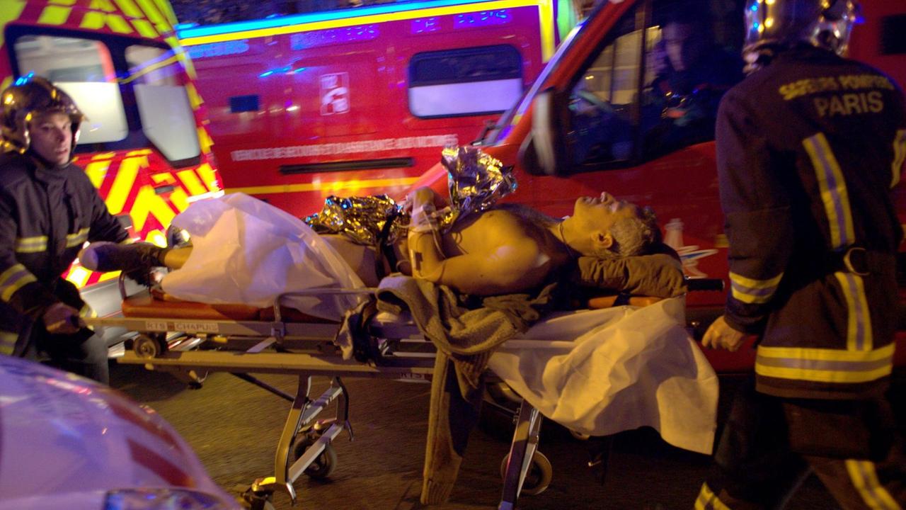 Zwei Sanitäter mit Helmen rollen den Verletzten an Krankenwagen vorbei. Der Mann ist unbekleidet und nur mit einer Goldfolie bedeckt. 