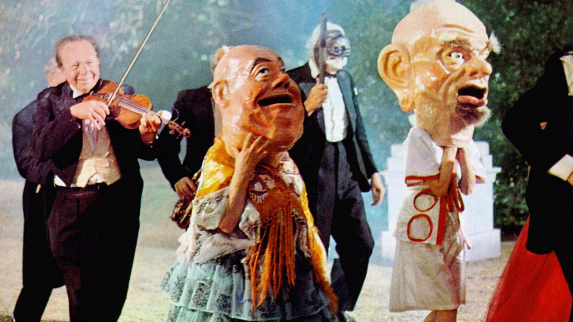 Szenenfoto aus dem Film Zardoz. Es schein eine Art Parade zu sein, links geht ein Geige-spielender Mann im Frack, neben ihm ebenso dunkel gekleidete Gestalten mit Blasinstrumenten und mit Masken, im Vordergrund rechts zwei große Figuren mit überdimensionalen Pappmaché-Köpfen