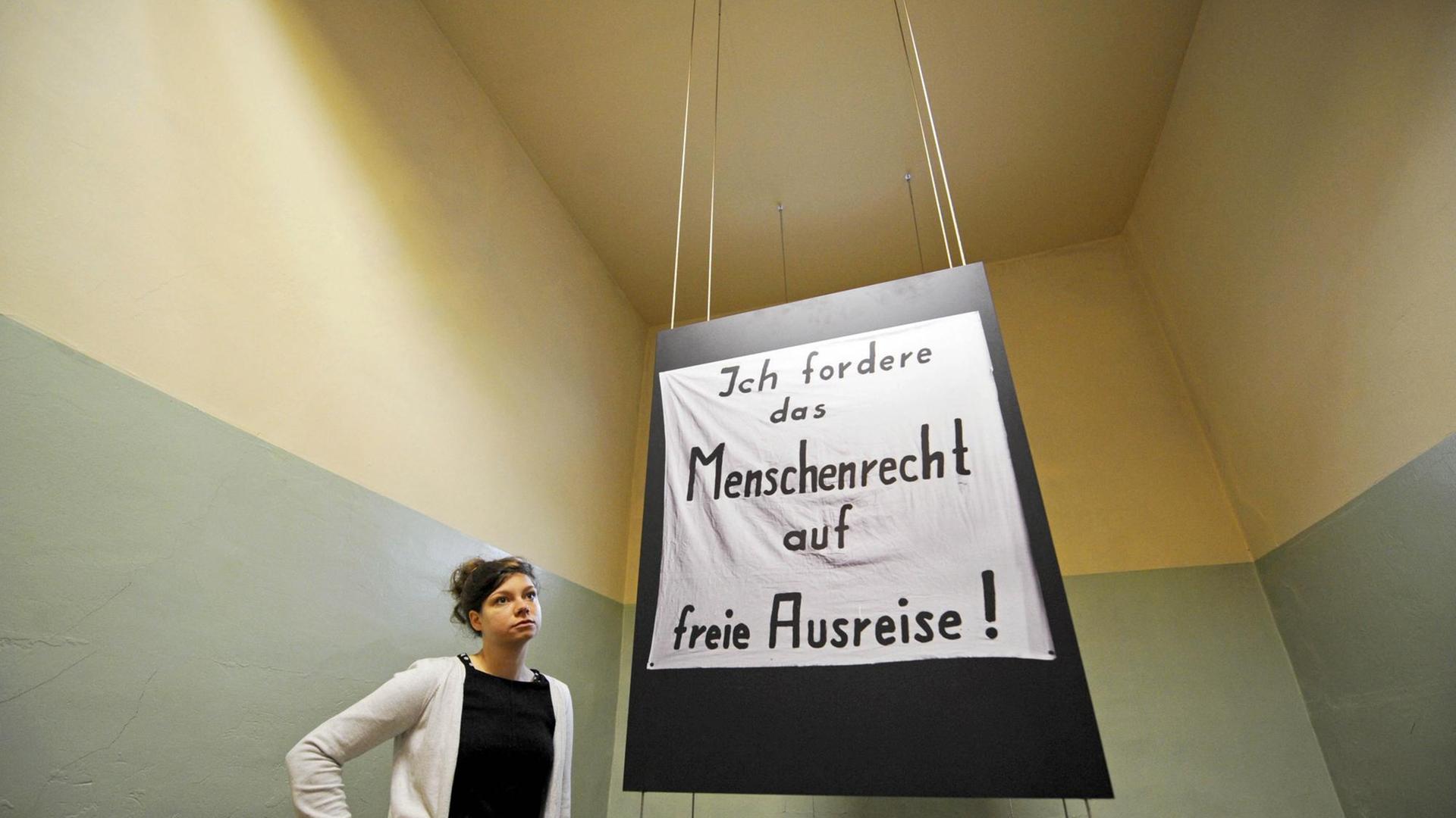 Eine Museumspädagogin steht in einer früheren Zelle des ehemaligen Stasigefängnis Andreasstraße in Erfurt, von der Decke hängt ein Plakat mit der Aufschrift "Ich fordere das Menschenrecht auf freie Ausreise!".