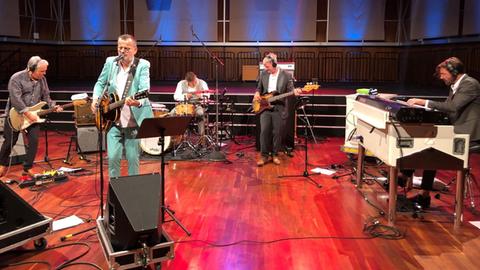 Fünf Musiker stehen in einem großen Saal mit verschiedenen Musikinstrumenten: in der Mitte der Sänger und Gitarrist Phillip Fankhauser in mint-grünem Anzug.