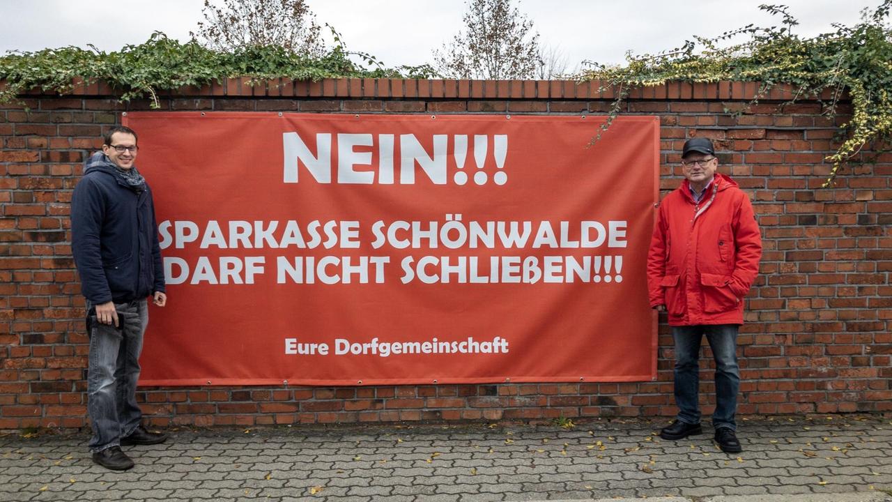 Grünen-Politiker Benjamin Raschke (links) und Bürgermeister Roland Gefreiter vor einem Plakat mit der Aufschrift: "Sparkasse Schönwalde darf nicht schließen"
