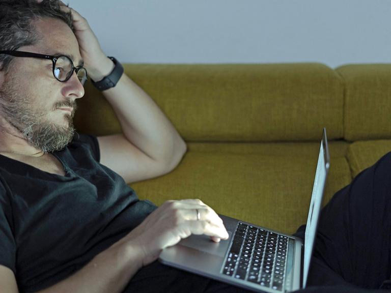Ein Mann mit Brille liegt auf dem Sofa und bedient einen Laptop.