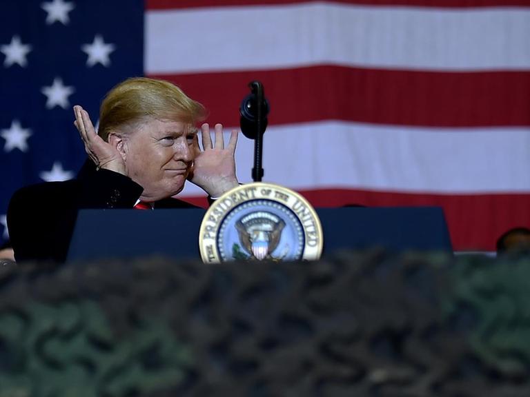 US-Präsident Donald Trump auf Kurzbesuch bei den US-Soldaten in Afghanistan. Er steht an einem Redepult und hält sich die Hände an die Ohren, als warte er auf eine Reaktion des Publikums.