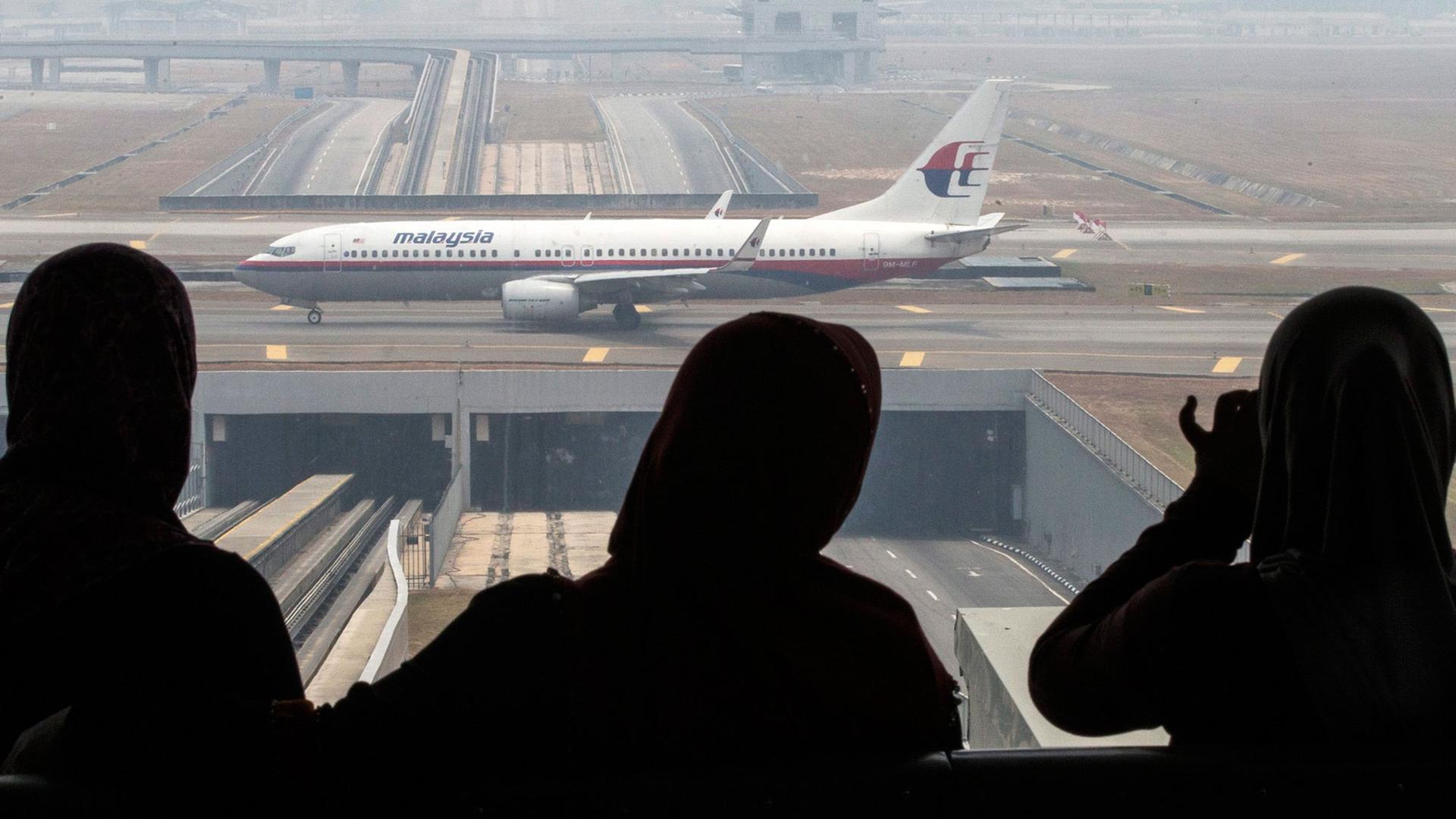 Drei Frauen blicken durch ein Fenster auf eine Boeing 777-200 von Malaysia Airlines.