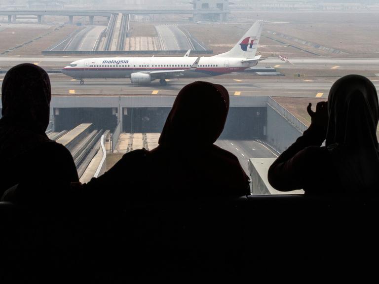 Drei Frauen blicken durch ein Fenster auf eine Boeing 777-200 von Malaysia Airlines.