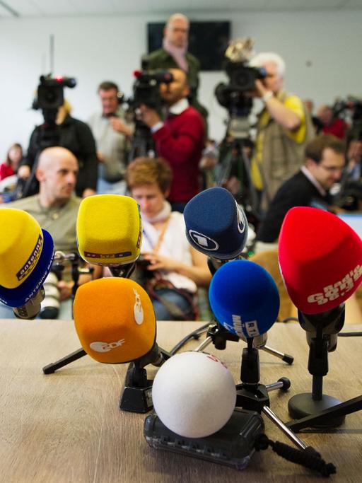 Auf einem Tisch stehen ein Dutzend Mikrofone, im Hintergrund sind Journalisten, zum Teil mit Kameras zu sehen.
