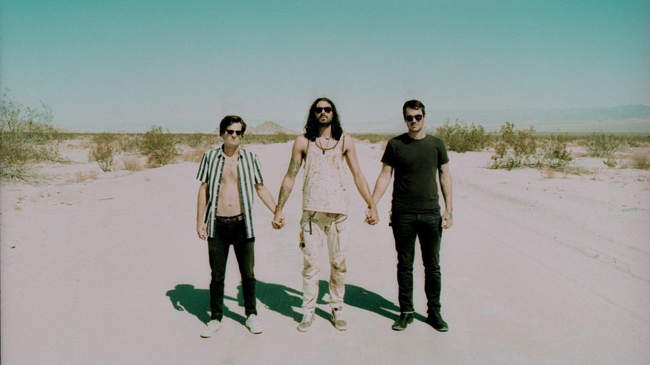 Drei Männer stehen in einer Wüste. Sie halten sich an den Händen und blicken in die Kamera.