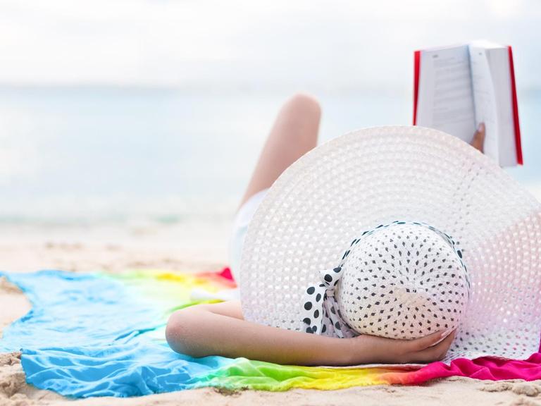 Eine Frau mit weißem Sonnenhut liegt am Strand und liest ein Buch. Ihr Handtuch strahlt in Regenbogenfarben.