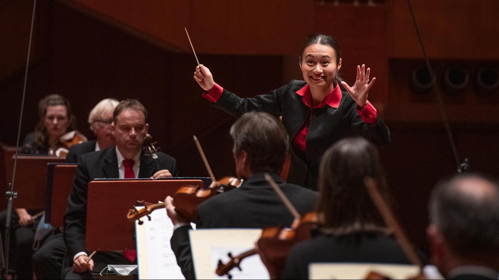 Eine junge Frau blickt freudestrahlend ins Orchester, das vor ihr sitzt. Sie hat die Hände zum Dirigat gehoben, in ihrer rechten Hand hält sie einen Dirigierstab hoch.